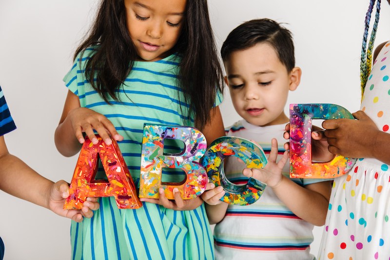 Large letter teacher appreciation crayon gift. Image is of four kids holding giant letters crayons in the shape of A, B, C, and D. Each custom crayon letter is made from recycled crayon in rainbow mix.