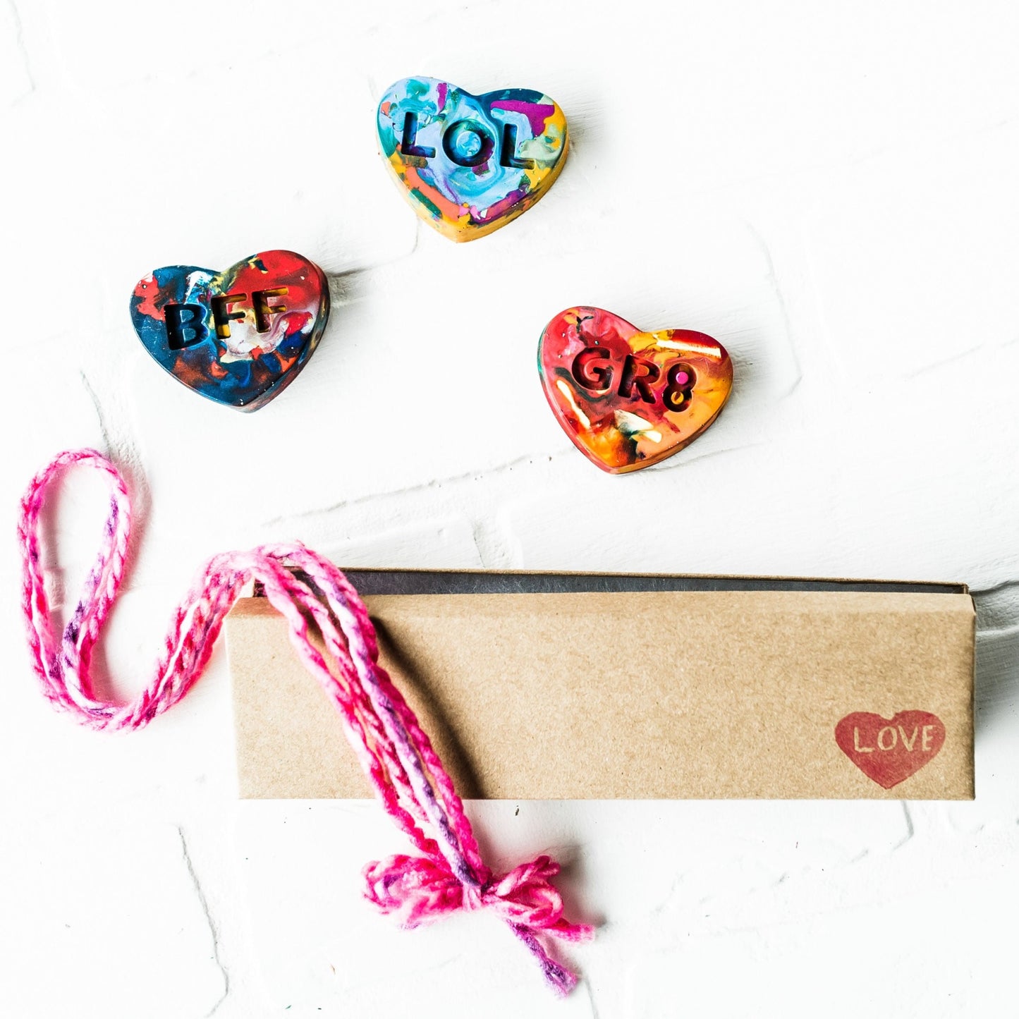 Three colorful conversation heart rainbow crayons from crayon shop Art 2 the Extreme are displayed on a white background. The kraft gift box they come shipped in is displayed, closed with a red LOVE heart stamp in the lower right corner and colorful pink yarn off to the left side. This unique Valentine's Day gift for kids comes ready to gift!