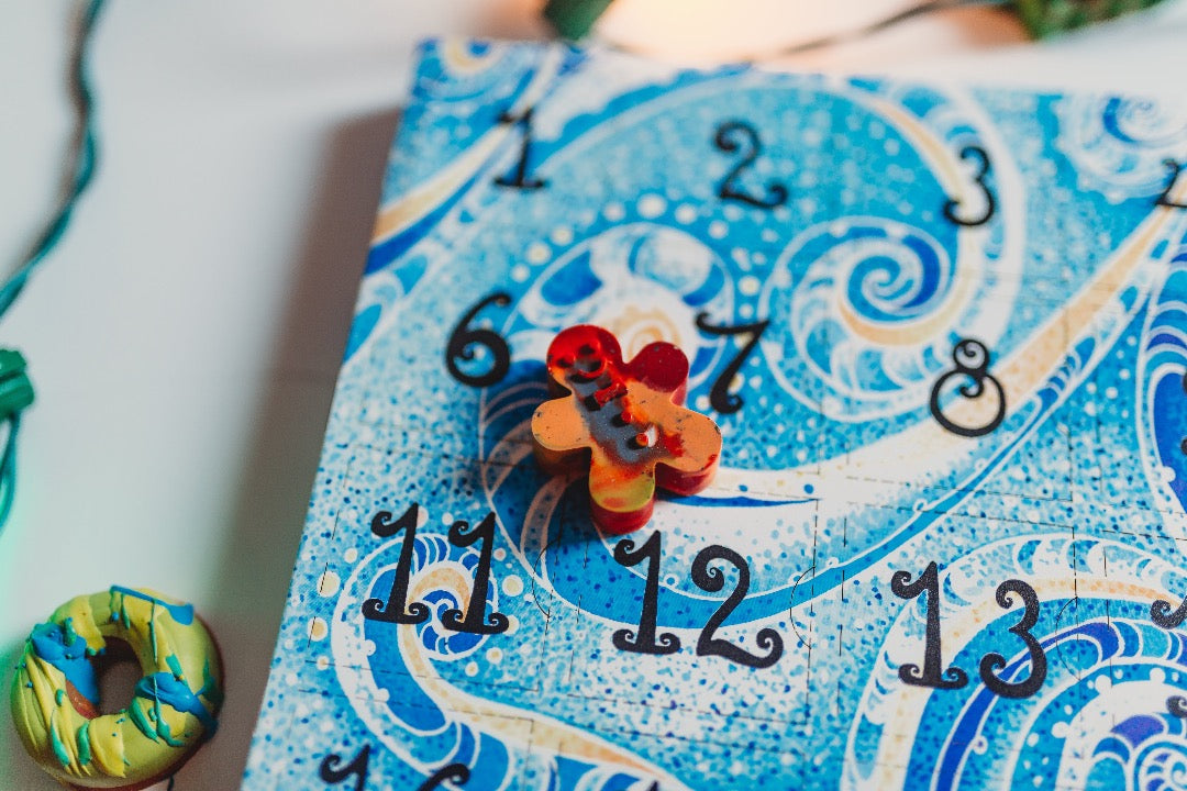 Closeup of blue paper crayon advent calendar for kids. A mini gingerbread Christmas crayon sit on top of a paper advent calendar for kids. The boxes 1-3, 6-8, and 11-12 show.