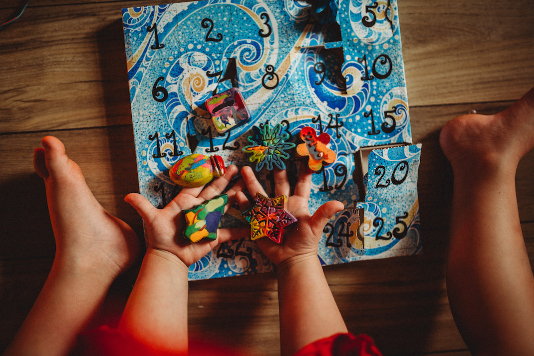 Child in red Christmas dress holds a handful of mini rainbow crayon shapes that came out of a paper Advent calendar, sitting on the floor in front of him. The advent calendar has a blue swirl pattern with the paper door numbers printed in black. Crayon Advent calendar is sitting on a wooden floor.