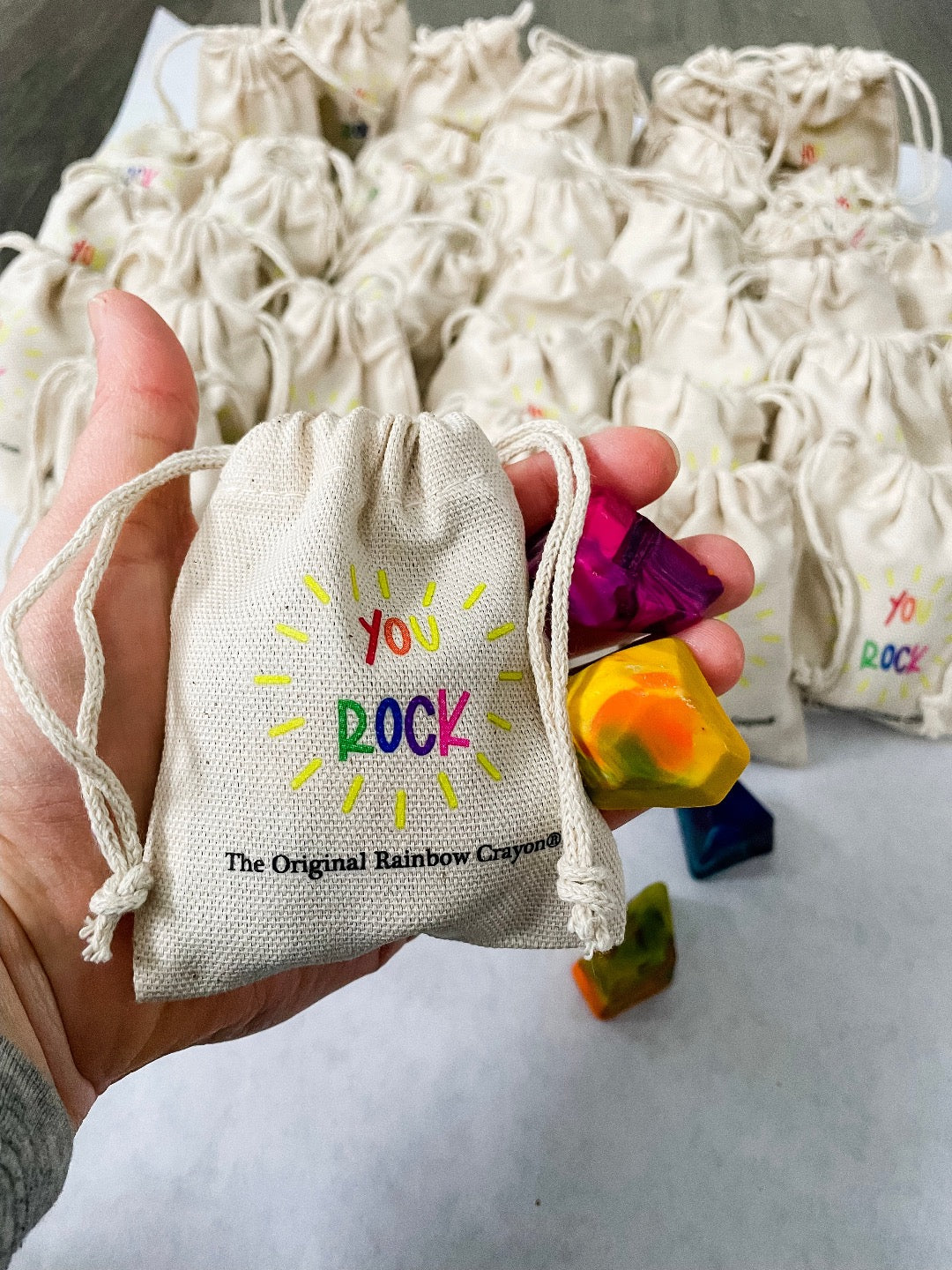 Adult hand holding the mini beige drawstring bag that has the test YOU ROCK in rainbow letters . The Original Rainbow Crayon logo is much smaller underneath in black text. Hand is also showing a yellow and pink mini crayon rock for scale. Bag fits perfectly in the hand of an adult .