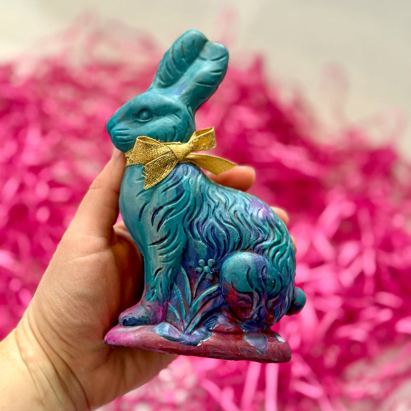 Hand holding a colorful Easter Bunny crayon for kids, handcrafted in a mix of blue, purple, and pink hues with a gold ribbon tied around its neck. This unique handmade crayon is shaped like a vintage-style rabbit and makes a perfect Easter basket stuffer or spring art gift for children. The background features bright pink Easter grass, adding a festive touch.