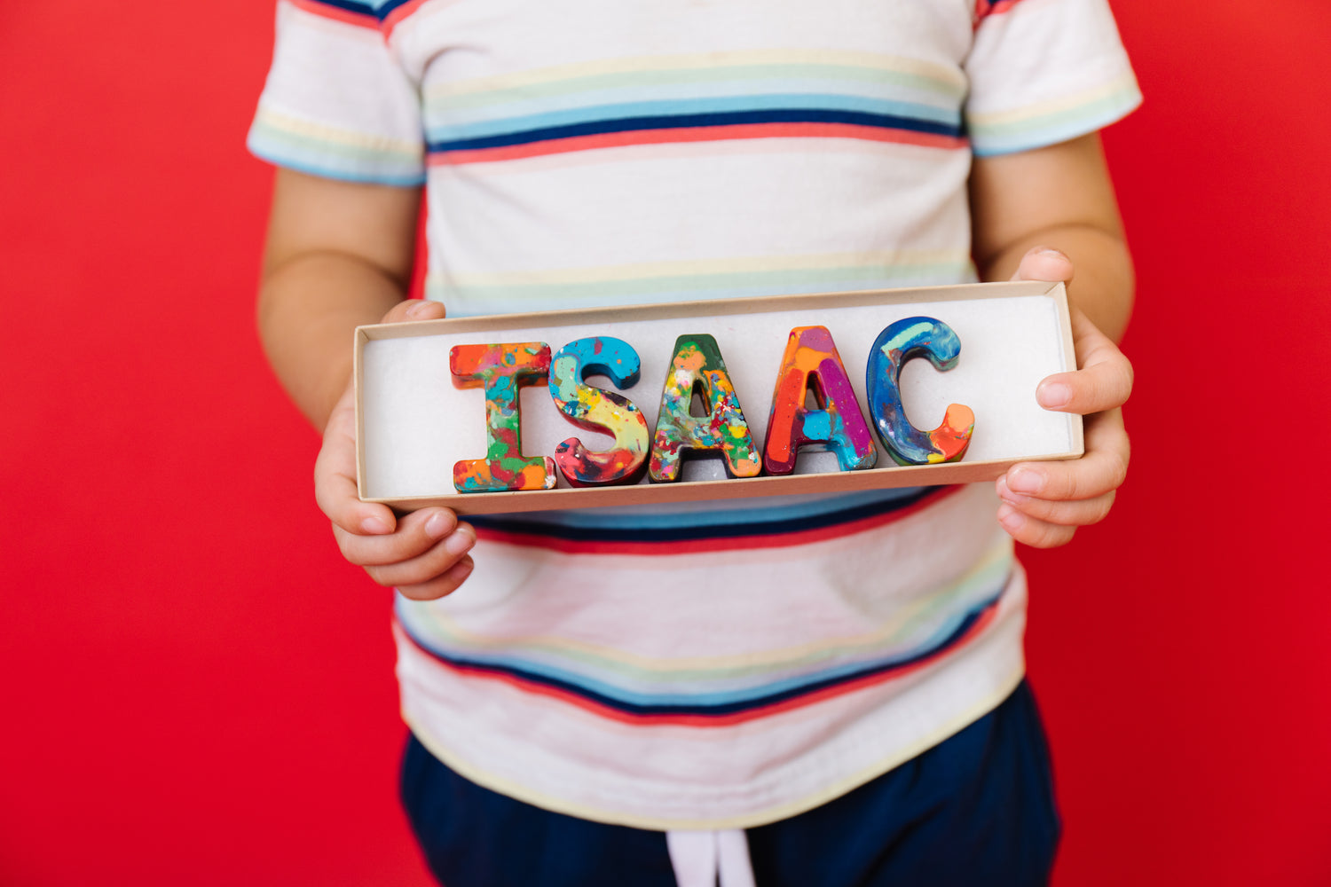Image of boy holding the name Isaac in rainbow crayon colors. Background is red and each individual letter crayon is made with rainbow colors. Create amazing crayon art with melted crayons. Rainbow crayons are the best-selling birthday gift for kids and stocking stuffer for kids. Crayon gifts for kids are perfect for a crayon artist, master crayon artist, or as toddler crayons. Tienda Rainbow 