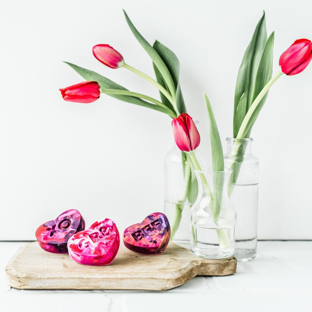 Three conversation heart crayons in pink/purple/red/peach combo. Crayons are propped on a charcuterie board as a photo prop with a vase of red roses next to them. The table and background are white. Multicolored heart crayons from Art 2 the Extreme make cute class valentines!