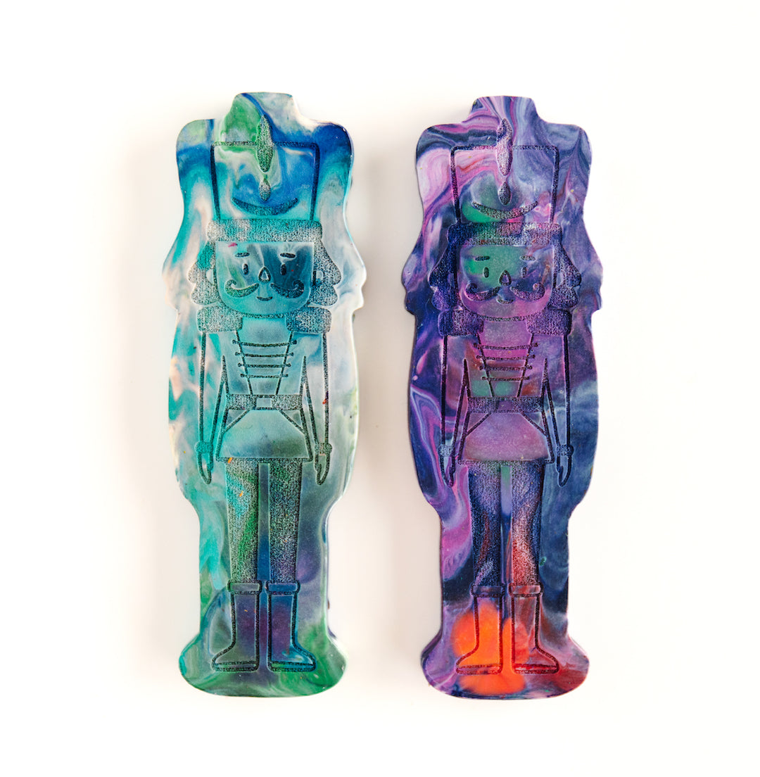 Two Nutcracker gift crayons from Art 2 the Extreme, shaped like a classic nutcracker figure with vibrant multicolors swirled throughout in cool colors such as blue, teal, purple, and green on a white background. The crayon features intricate detailing, including a tall hat, mustache, and uniform-like design, making it a festive and creative holiday gift for children who love coloring.