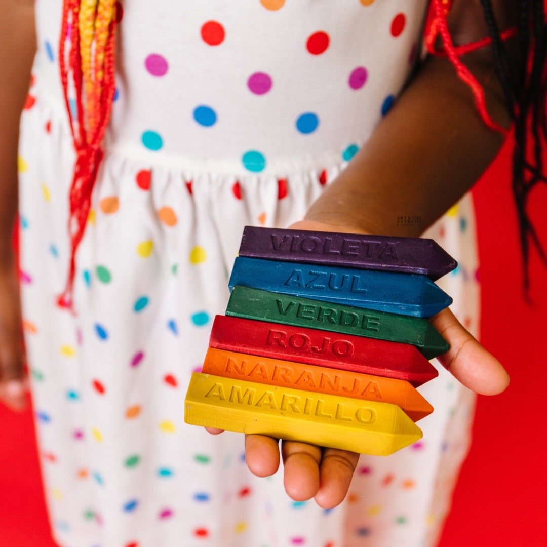 Girl wearing polkadot dress holds 6 Spanish learning crayons in her hand. Each Original Rainbow Crayon® Stix has the Spanish color name engraved on them. 9 oversized Spanish crayons are included.