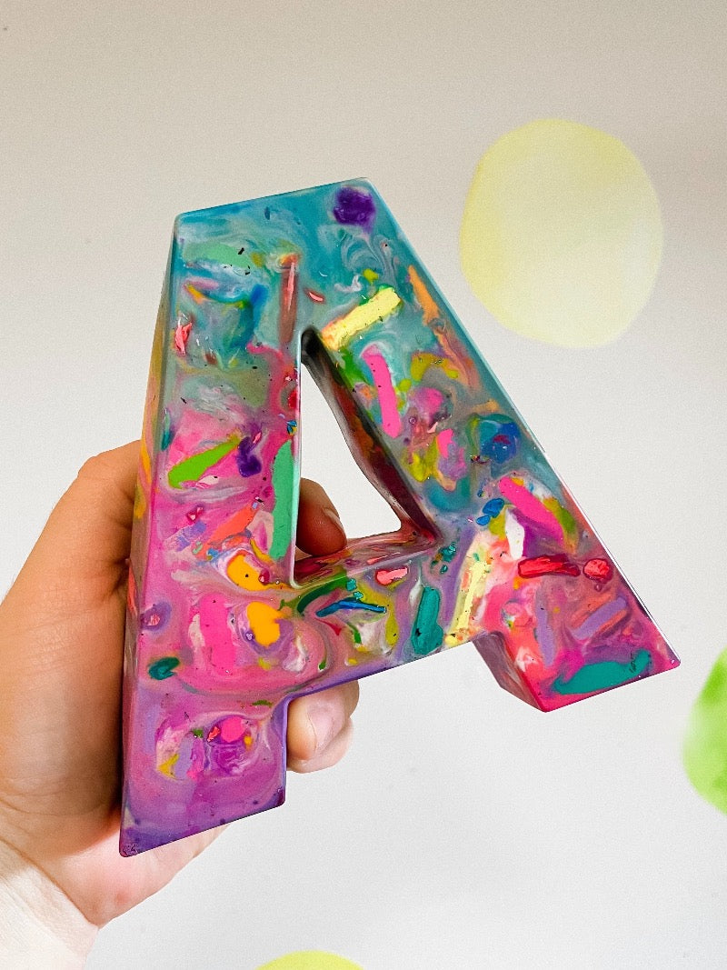 Huge letter A. Image is of an adult hand holding up a Crayon letter A on a white background. Crayon colors are in a mix of pastel pinks and teals. This oversized crayon is in the shape of a large letter A. This rainbow crayon from crayon shop, Art 2 the Extreme, has been created with pastel colored recycled crayons. This unique gift for kids is larger than an adult hand and perfect for back to school gift ideas or stocking stuffers for kids. Pair with a kids paper pad!
