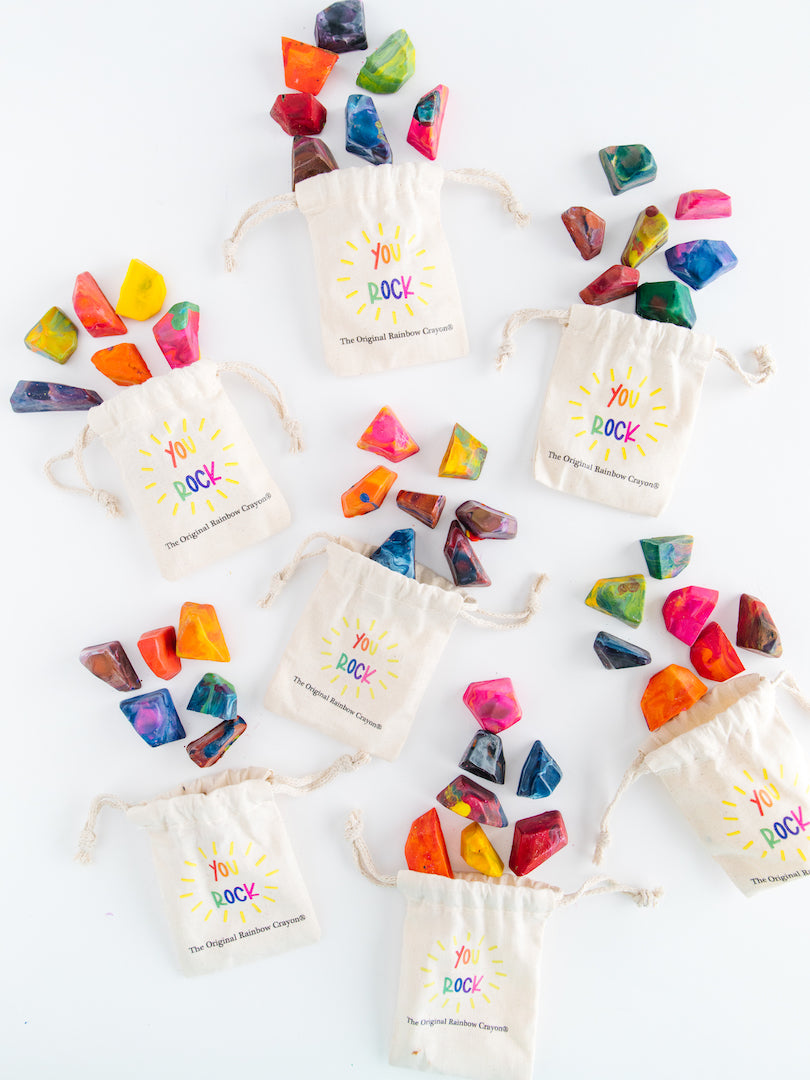 Colorful rock crayons. 7 Mini bags with each bag showing 7 mini rainbow rock crayons. Each off-white drawstring bag says YOU ROCK and Original Rainbow Crayon logo on it. Crayon gift sets are displayed on a white background so the bright colored crayons almost glow!