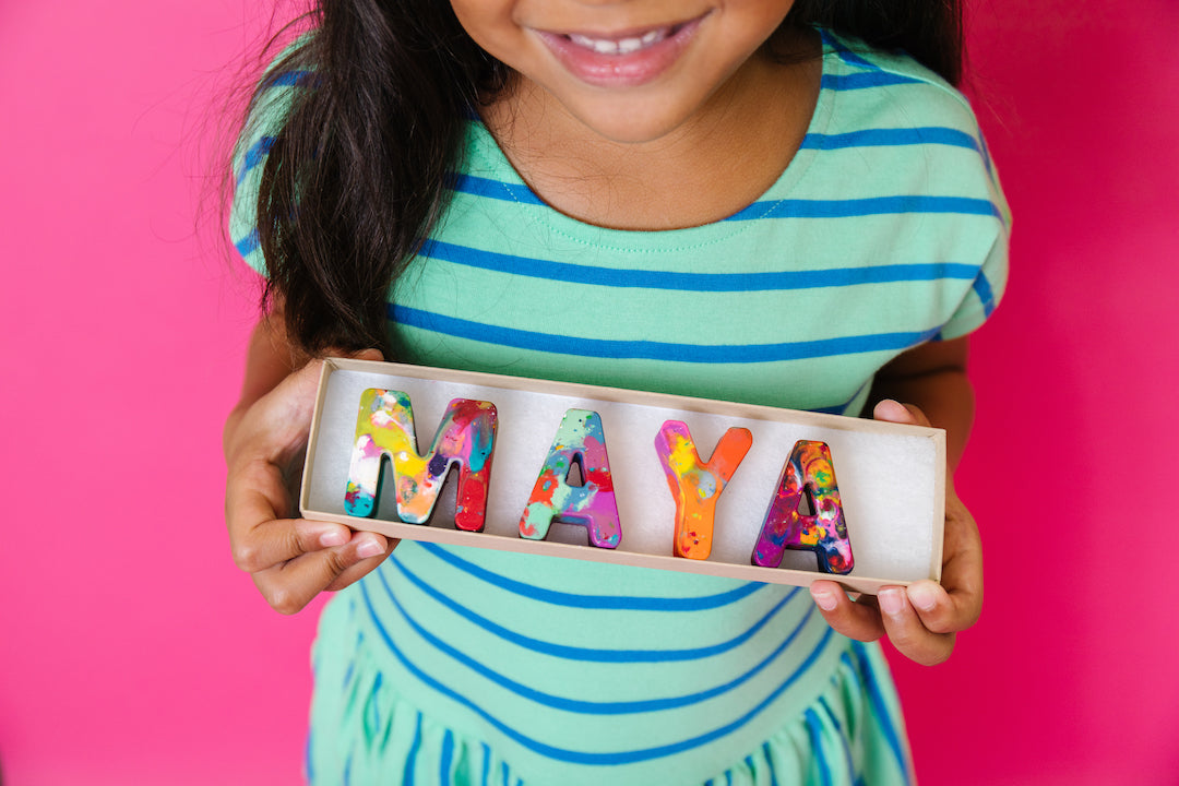 Girl holding a personalized name crayon set that spells MAYA in  rainbow crayon colors. The background is pink. This customized crayon gift is a perfect easter gift for little girls. 