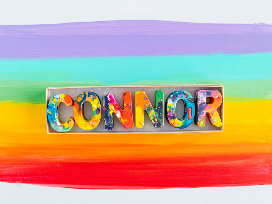 Name crayon set in gift box that spells the name CONNOR in colorful recycled crayon letters. Image of custom crayons is on a white background with painted rainbow lines in the colors of purple at the top, teal, green, yellow, orange, and red at the bottom. 