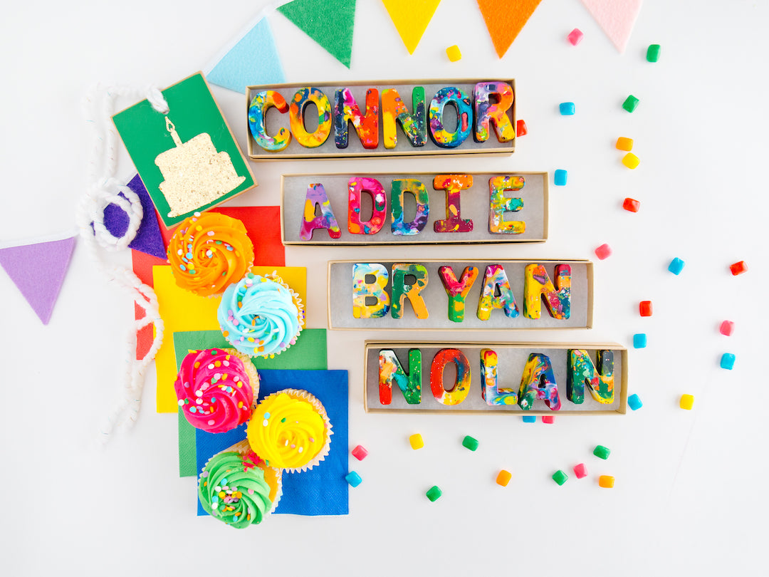 Custom crayons that spell out the names CONNOR, ADDIE, BRYAN and NOLAN in kraft colored gift boxes that are 8"x2". Personalized crayon name sets are on a white background with confetti pieces, birthday banner, and birthday cupcakes as birthday themedphoto props. 