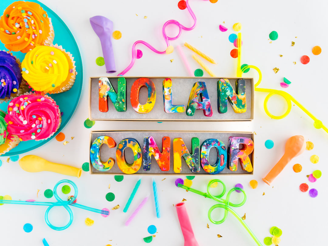 2 Personalized birthday gift crayon names that spell the name NOLAN and CONNOR in bright, colorful rainbow colors. The oversized letter crayons look tie dye with colorful flecks mixed in. Background is white with confetti , balloons, and rainbow cupcakes as photo props.