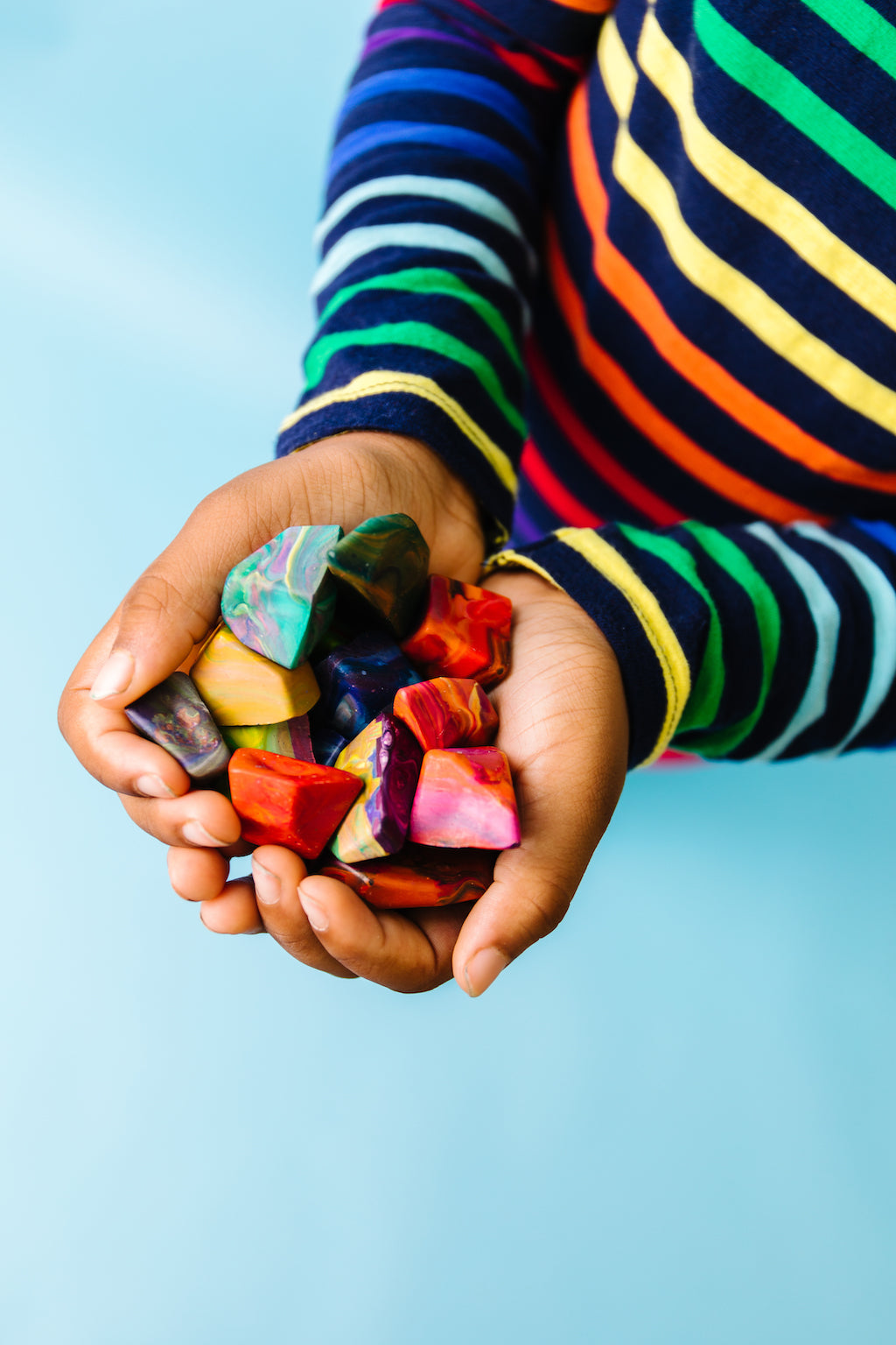 Child holding a handful of multicolored rock crayons in different hues. Handful has about 15 rainbow rock crayons on a teal blue background.