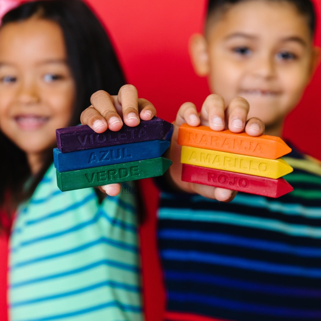 Learn colors in Spanish with Original Rainbow Crayon Spanish color crayon set. Image is of two kids each holding out three solid colored, oversized crayons with the Spanish names engraved on them.