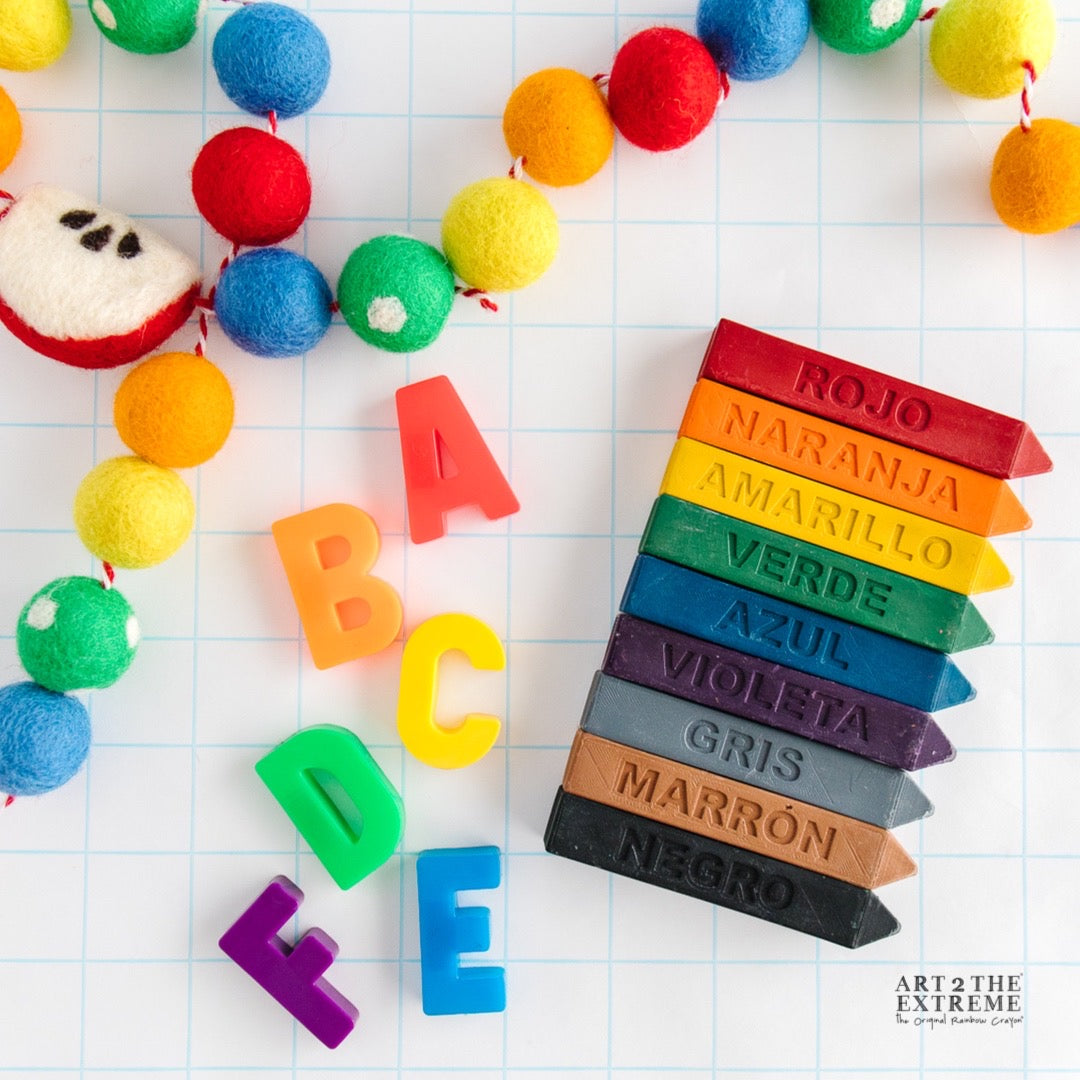 9 spanish colored crayons are on a white background with ABC magnets surrounding them. These crayon gifts for kids are a great way to learn the colors in Spanish and a perfect classroom gift.