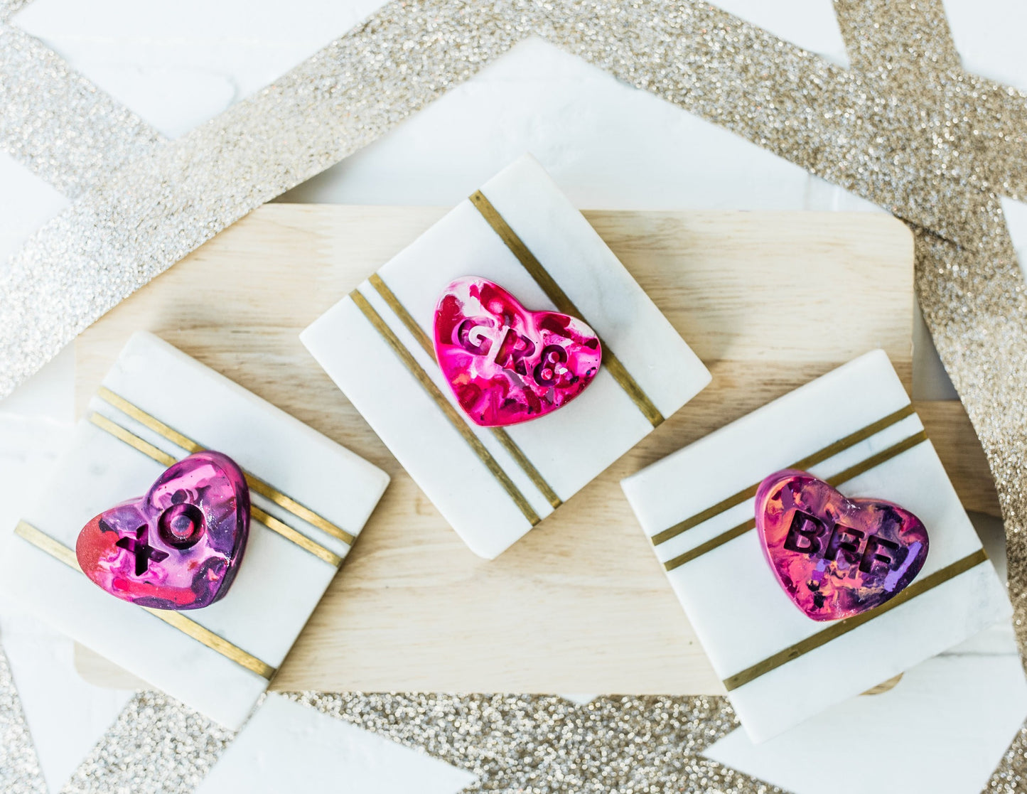 Three multi-colored conversation heart valentine crayons for kids are displayed on a white and gold background with a wooden charcuterie board as a photo prop. Rainbow crayons displayed are a pink/purple/red mixture for Valentine's Day.
