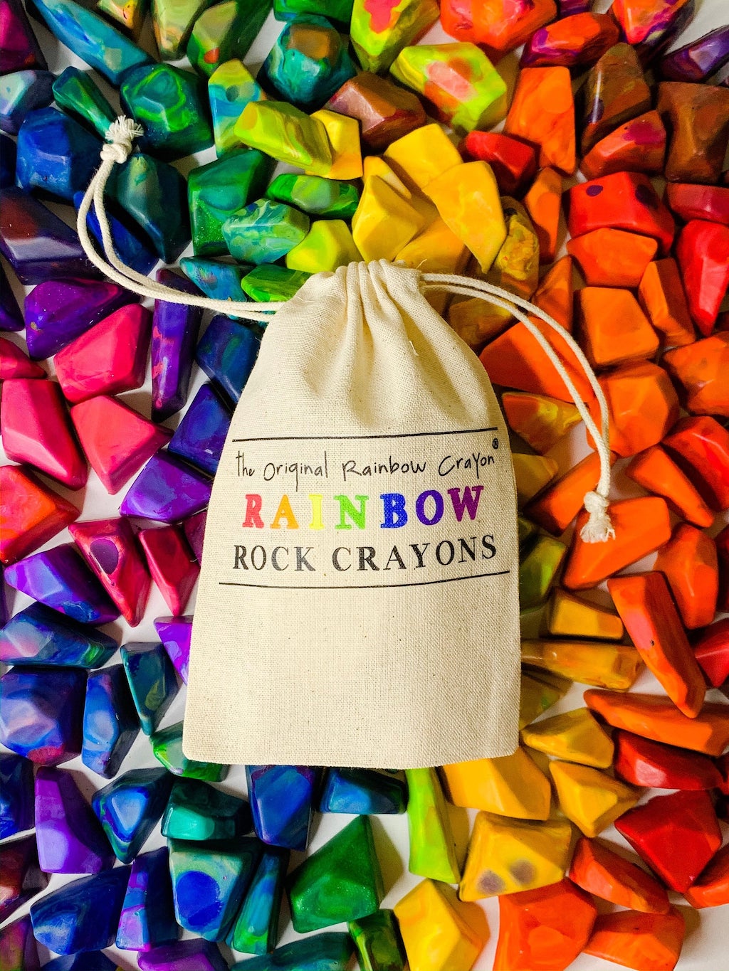 A canvas bag labeled "The Original Rainbow Crayon® - Rainbow Rock Crayons" is surrounded by colorful, gem-shaped crayon rocks arranged in a rainbow pattern