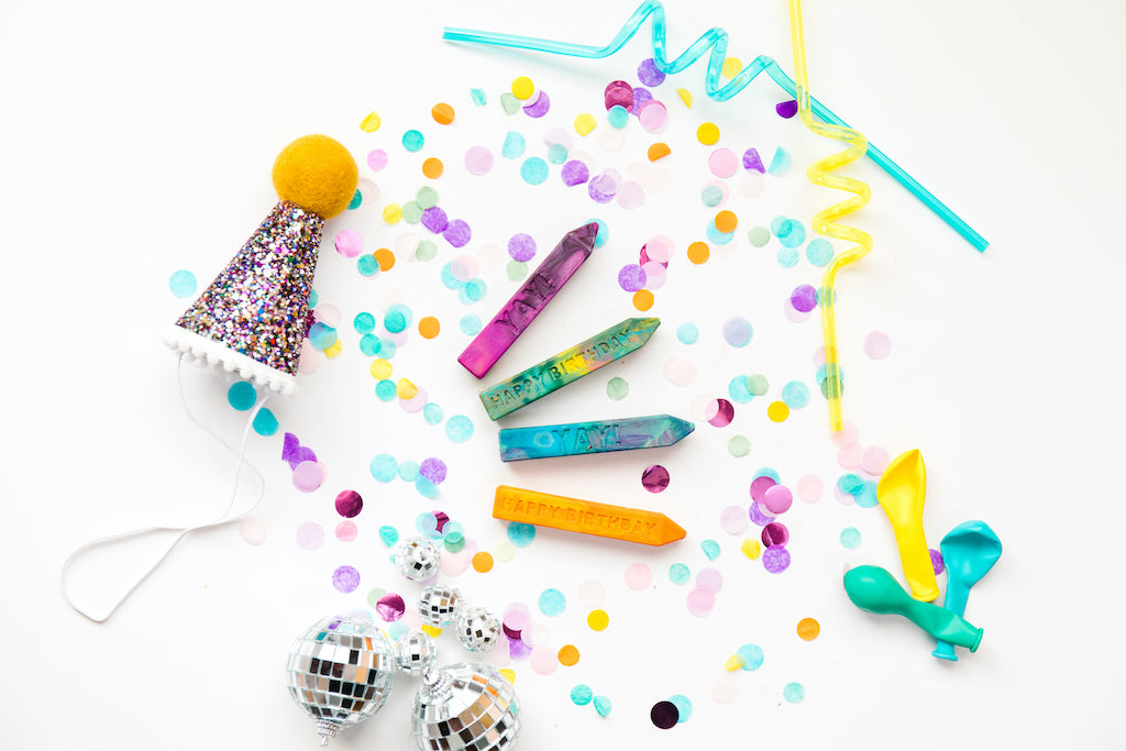 Four Crayon Stix® in colors purple, green, blue, and yellow I a white background with confetti. 2 birthday child crayon that say yay and 2 birthday child crayon that says Happy Birthday. Photo props of a tiny birthday hat, disco balls, balloons, and two curly straw favors are also in the photo.