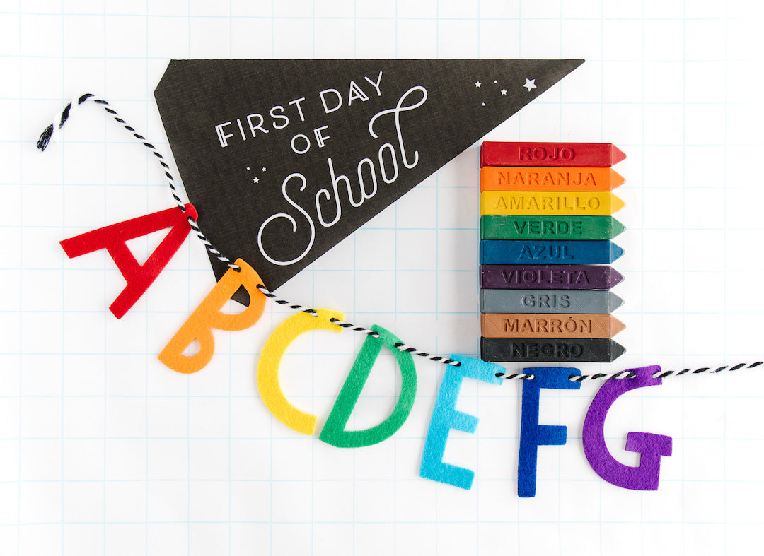 Nine solid colored rainbow crayon sticks with Spanish crayon names on a white background with a first day of school banner flag and ABC garland as photo props.