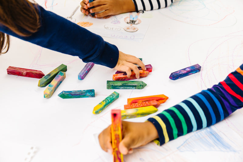 Children using colorful handmade rainbow crayons to draw together on paper, showcasing creative play and crayon gifts for kids during a group art activity.