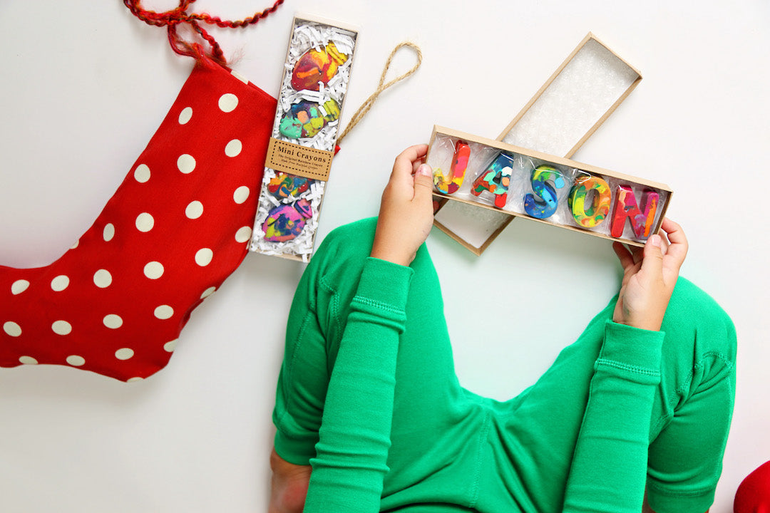 Child in matching green Christmas pajamas opening a crayon name set that says JASON in rainbow crayon letters.