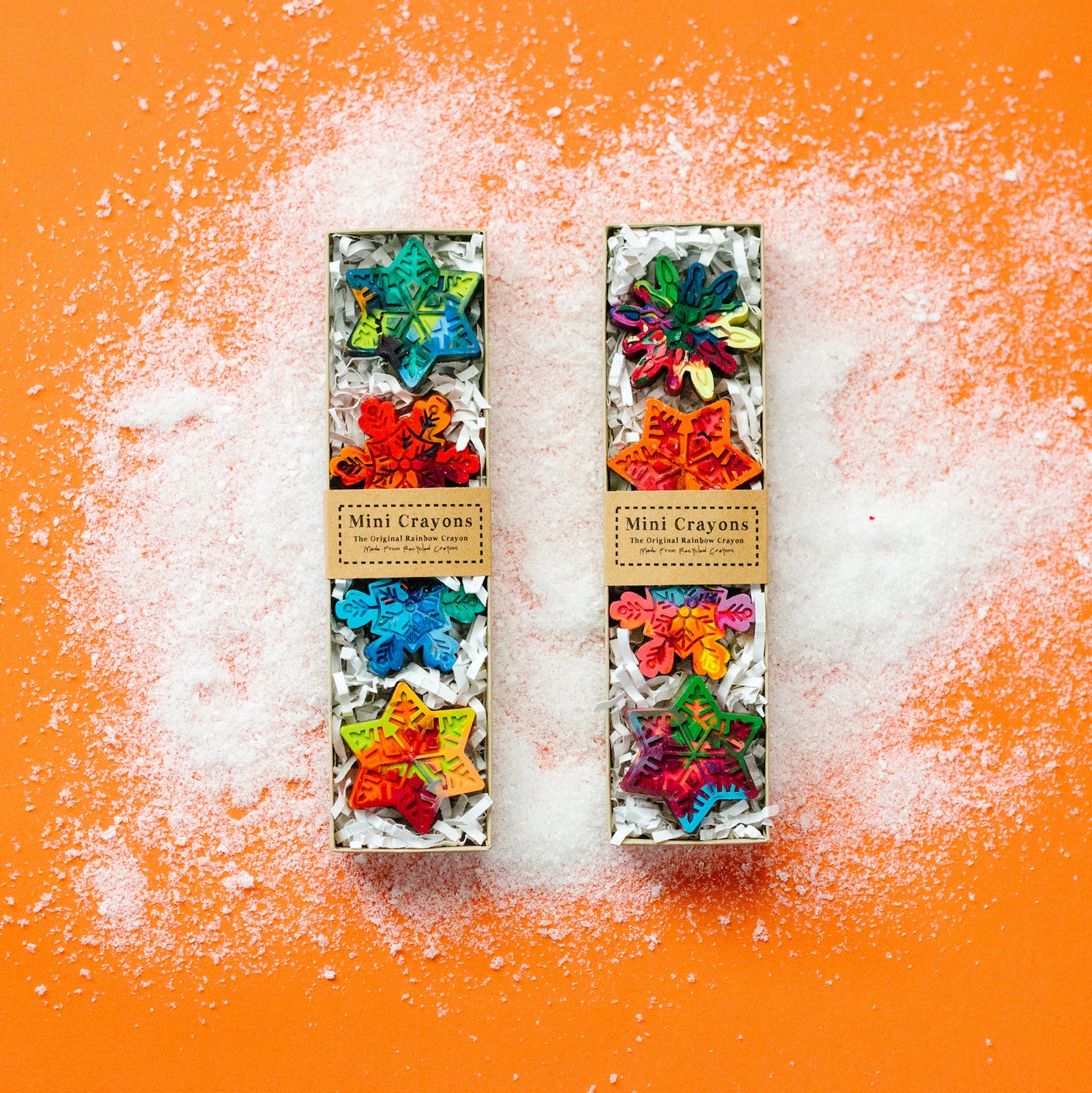 This image shows two festive Mini Snowflake Crayon gift boxes from Art 2 the Extreme. Each clear-top box contains four vibrant, multicolored snowflake-shaped crayons, nestled in white shredded paper, creating a winter holiday theme.
