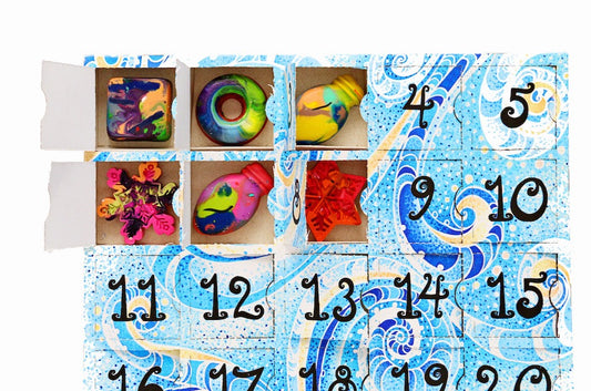 Kids advent calendar filled with crayons from handmade crayon shop, Art 2 the Extreme. A paper advent calendar with 6 doors punched open revealing mini rainbow crayon shapes sits on a white background. The crayon Advent calendar for kids has a blue , teal, and light yellow design with numbered boxes. 