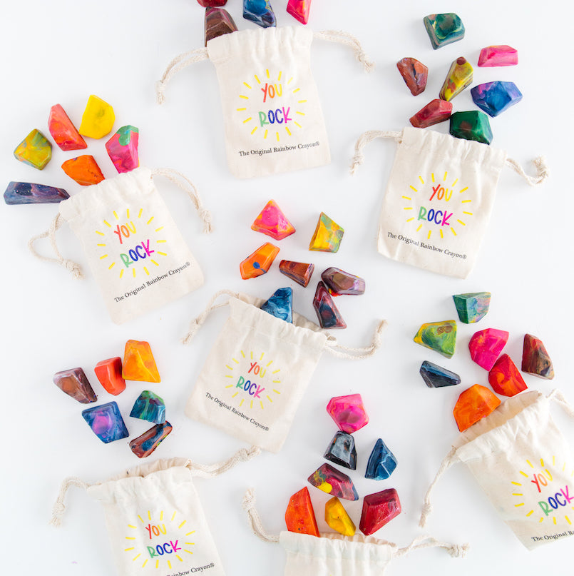 Seven bags of rainbow rock crayon rock favors. This crayon designs on the front of the bag say "You ROCK' in rainbow letters with the Original Rainbow Crayon logo underneath. Each off-white bag of crayon rocks have 7 min rock crayons laid out for display on a white background. This image is from photographer Qiana K Photography and not an ai artwork or ai art generator art. Everything is handmade by crayon artist, Nicole Lewis.