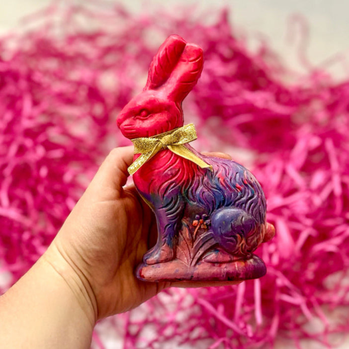 Hand holding a colorful Easter Bunny crayon for kids, handcrafted in a mix of pink and purple color hues with a gold ribbon tied around its neck. This unique handmade crayon is shaped like a vintage-style rabbit and makes a perfect Easter basket stuffer or spring art gift for children. The background features bright pink Easter grass, adding a festive touch.