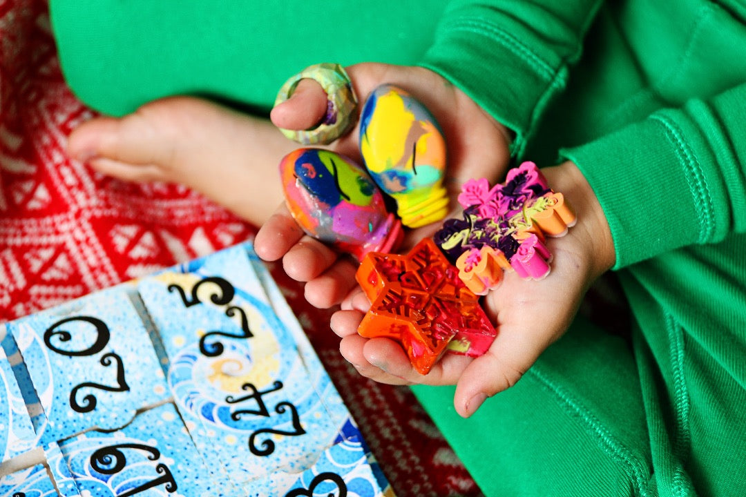 Child in green Christmas pajamas holds a handful of mini rainbow crayon shapes that came out of a paper Advent calendar, sitting on the floor in front of him. The advent calendar has a blue swirl pattern with the paper door numbers printed in black.