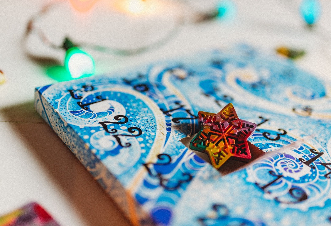 A blue crayon Advent calendar for kids sits on a white background with Christmas lights out of focus in the background. In box number 18, a mini Snowflake Original Rainbow Crayon is revealed with the paper calendar door open. Advent calendar is with a blue swirl design with black numbering on each of the punch out Advent Calendar doors.