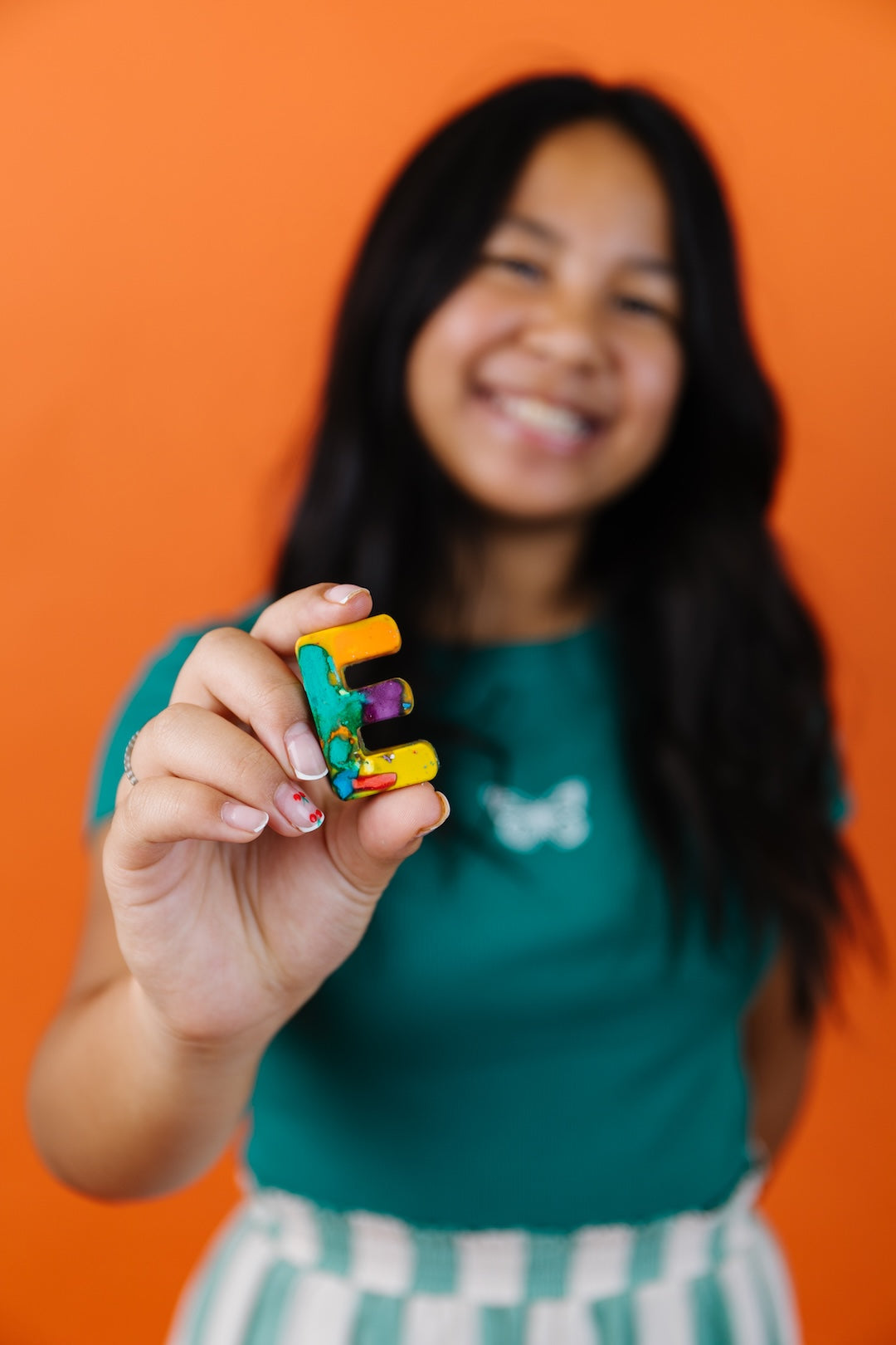 A smiling child holding a small, colorful, three-dimensional letter 'E' rainbow crayon against a solid orange background.