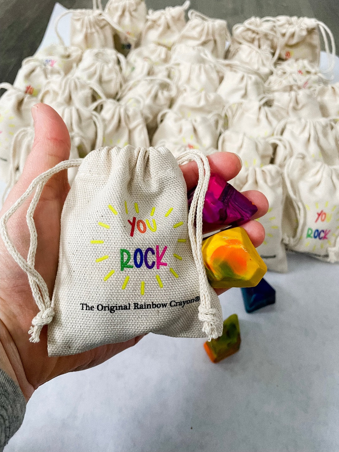 Adult hand holding  the mini beige drawstring bag that has the test YOU ROCK in rainbow letters . The Original Rainbow Crayon logo is much smaller underneath in black text. Hand is also showing a yellow and pink mini crayon rock for scale. Bag fits perfectly in the hand of an adult . 