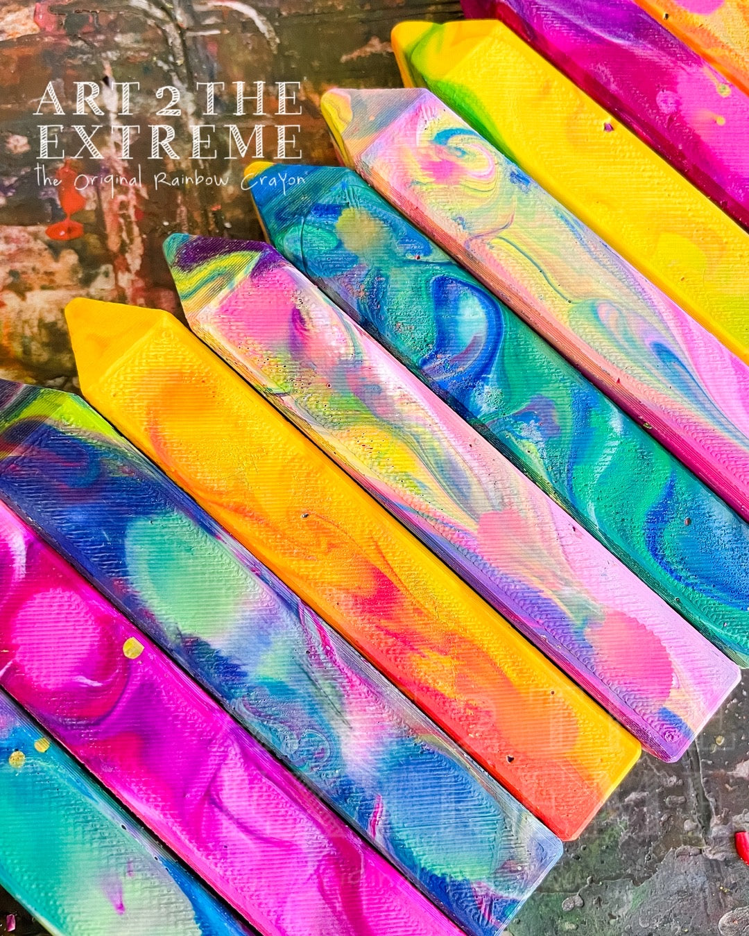 10 Individual Rainbow Crayon Stix that are multicolored, swirled crayons on a table.  Actual rainbow crayon gift product from Art 2 the Extreme will be shrink-wrapped in a pack of 4. 
