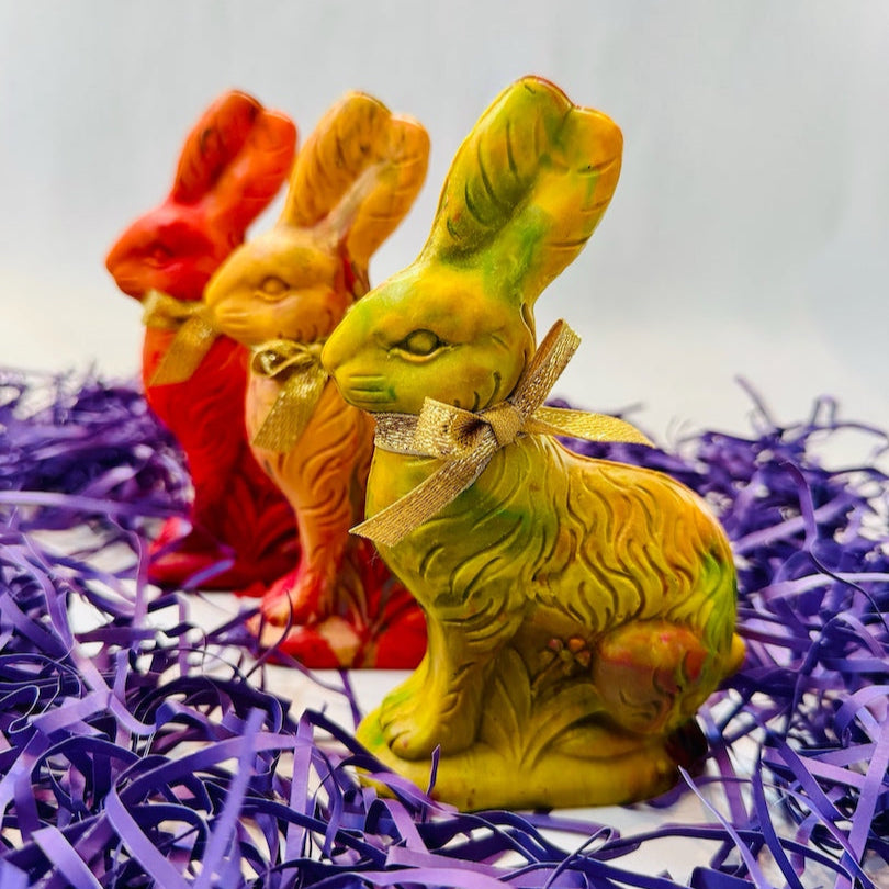 Three Easter Bunny crayons for kids in vibrant yellow, golden yellow, and orange hues, each adorned with a gold ribbon. These handmade bunny-shaped crayons are perfect for Easter basket gifts, party favors, and spring art activities. The colorful, vintage-style rabbits are displayed on a festive background with purple Easter grass, making them a goof gift for Easter or cute family Easter gifts.