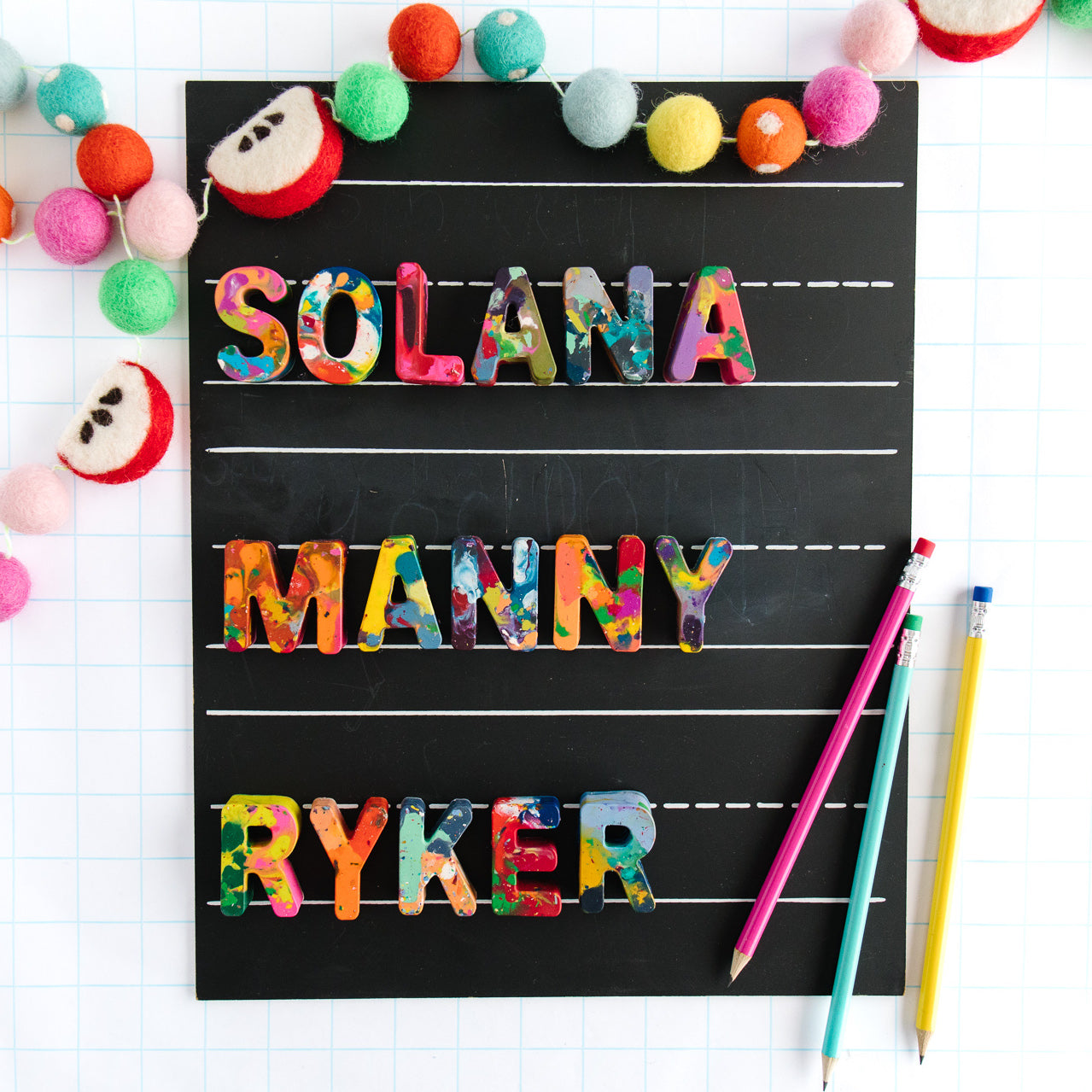 Back to school gift guide gift for kids. Three rainbow colored crayon names are displayed on a black chalkboard and a back to school themed apple garland borders the chalkboard as a photo prop. Rainbow Crayon names spell SOLANA, MANNY, and RYKER. 