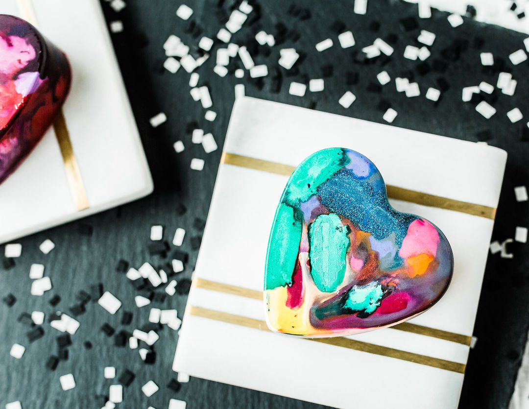 Close up of a colorful jumbo crayon heart that is mostly teal, pink, orange, with sparkle glitter crayons. This heart shaped crayon sits on a white tile with a gold line, which sits on top of black paper with white confetti used as a photo prop.