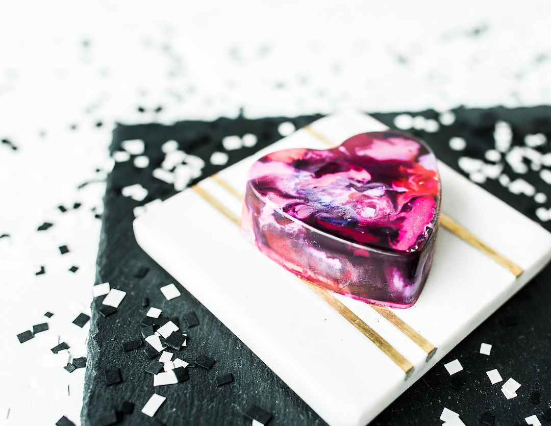 Close up of and a side view angle of a colorful jumbo crayon heart that is mostly pink, red, purple, violet, and white with sparkle glitter crayons. This heart shaped crayon sits on a white tile with a gold line, which sits on top of black paper with white confetti used as a photo prop.