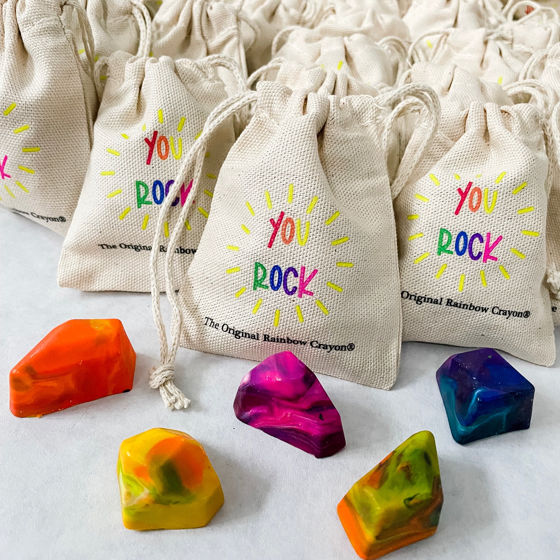 Several mini, beige drawstring bags with the text YOU ROCK is shown on a white paper. One bag is more in focus with 5 multicolored crayon rocks on top of the paper. The Original Rainbow Crayon logo is also shown in smaller, black letters below the rainbow YOU ROCK saying. 