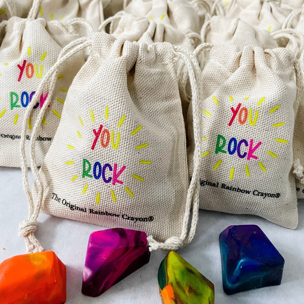Close up of several mini, beige drawstring bags with the text YOU ROCK is shown on a white paper. One bag is more in focus with 5 multicolored crayon rocks on top of the paper. The Original Rainbow Crayon logo is also shown in smaller, black letters below the rainbow YOU ROCK saying. 