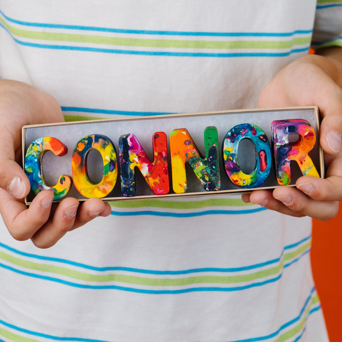 A person wearing a white striped shirt holds a rectangular box containing colorful, rainbow crayon letters spelling the name "CONNOR.".