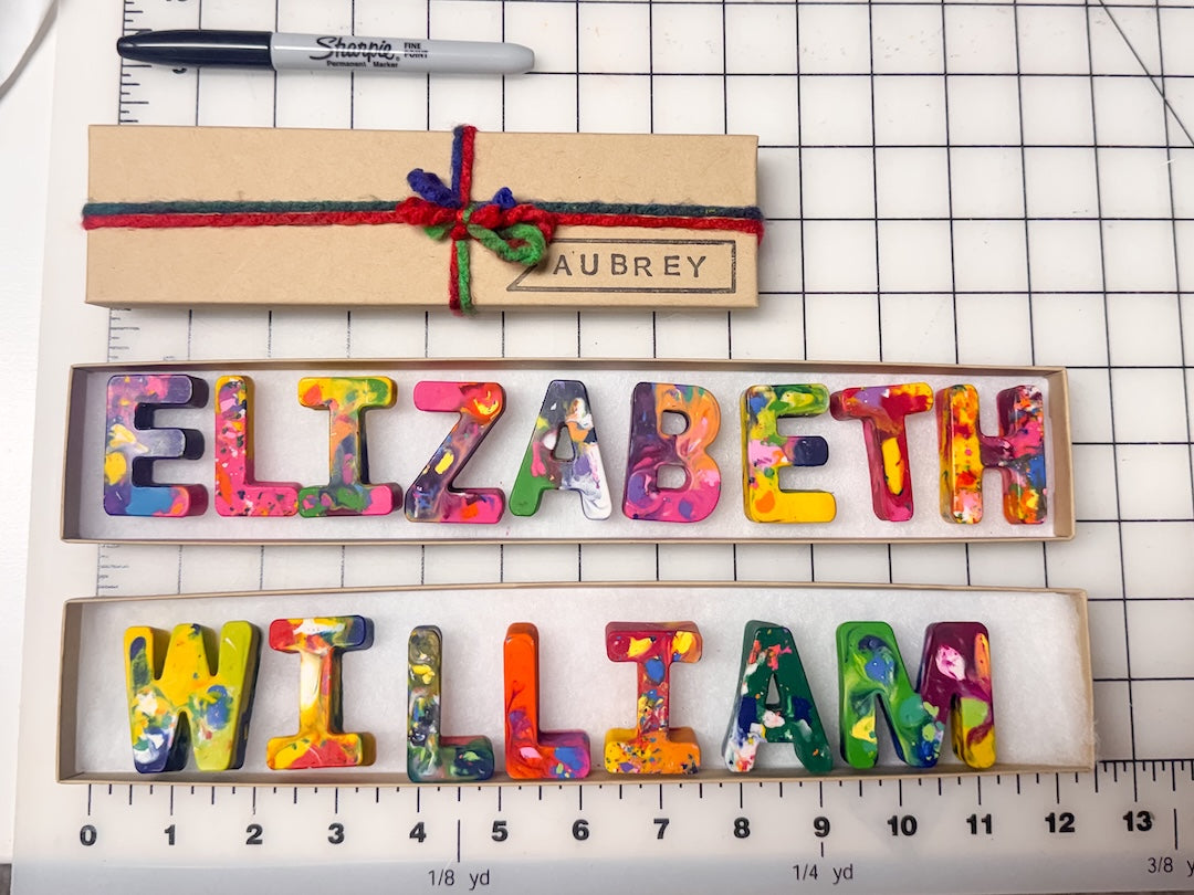 Personalized crayon name letters spelling 'ELIZABETH' and 'WILLIAM' in a colorful marbled pattern, displayed in their custom cardboard kraft gift boxes on a gridded cutting mat background.