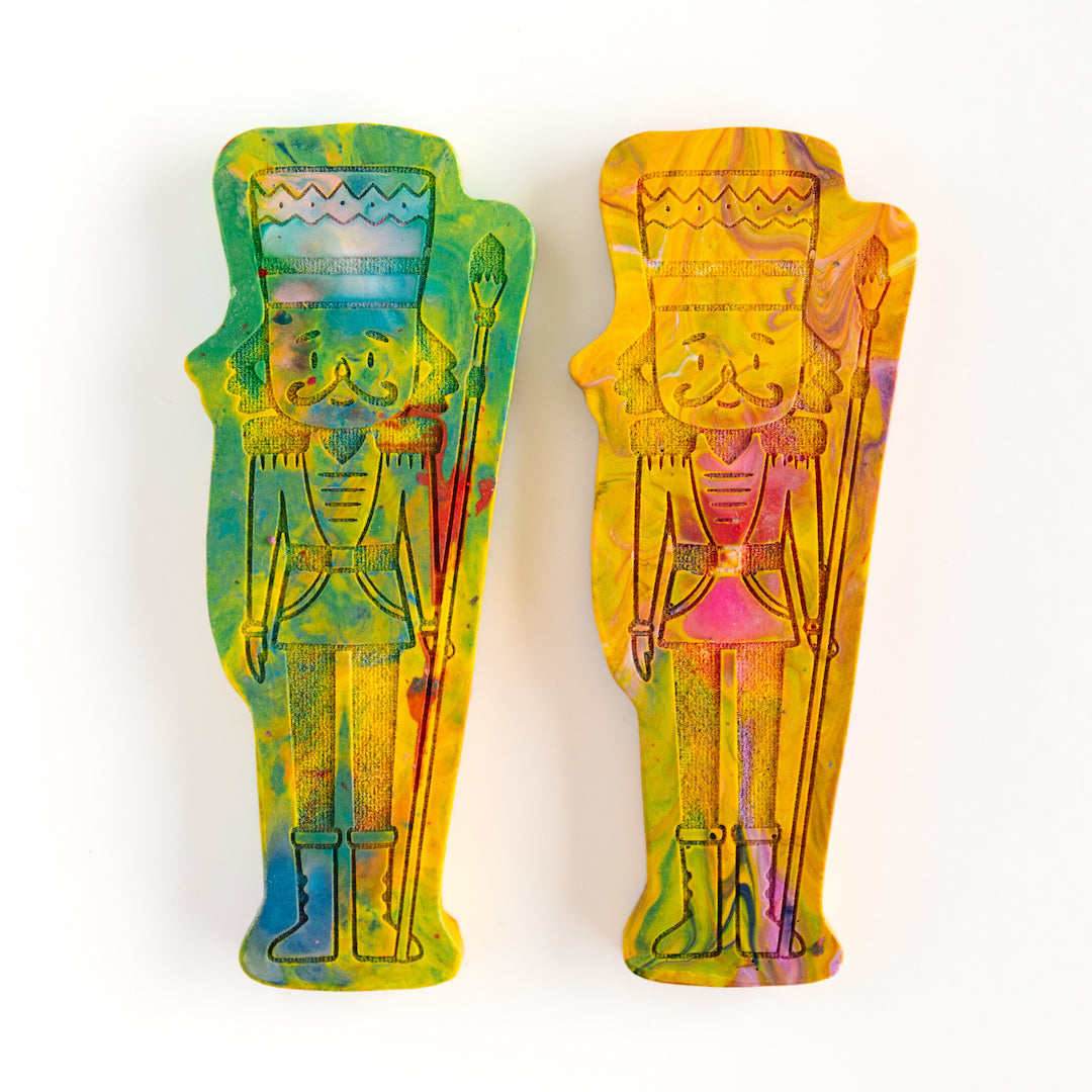 Two Nutcracker soldier crayons in green and yellow colors from Art 2 the Extreme, shaped like a classic nutcracker figure with vibrant multicolors swirled throughout. The crayon features intricate detailing, including a tall hat, mustache, and uniform-like design, making it a festive and creative holiday gift for children who love coloring.