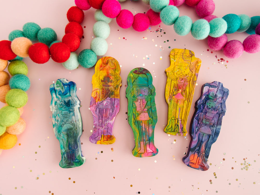 Five Nutcracker-shaped rainbow crayons from Art 2 the Extreme are displayed on a pink background, surrounded by colorful felt ball garlands in red, green, pink, and blue. Each nutcracker ballet gift ideas crayon showcases a unique blend of swirling colors, such as blue, yellow, green, and purple, with intricate Nutcracker details. The scene is sprinkled with glitter, adding a festive touch, making these crayons ideal for Christmas gifts, Nutcracker-themed crafts, and stocking stuffers for kids.