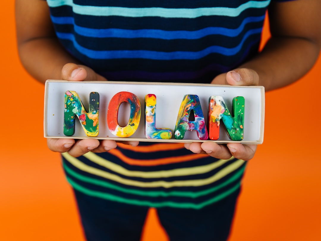 A child in a blue and black striped shirt holds a white tray containing colorful rainbow crayon letters spelling 'NOLAN' against a bright orange background.