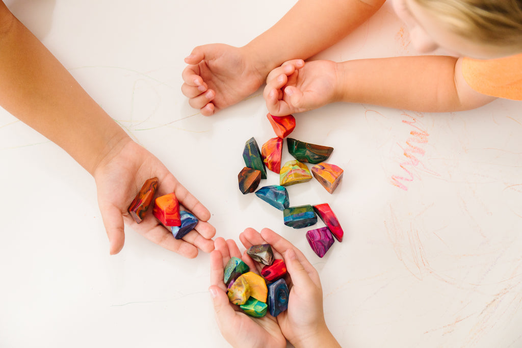 Two children arms and hands shown on a white background (paper). An older child holds 9 multicolored rock crayons in their hands while the other colors with  11 rock crayons spread onto of the white background. A red scribble is shown on the white paper. 