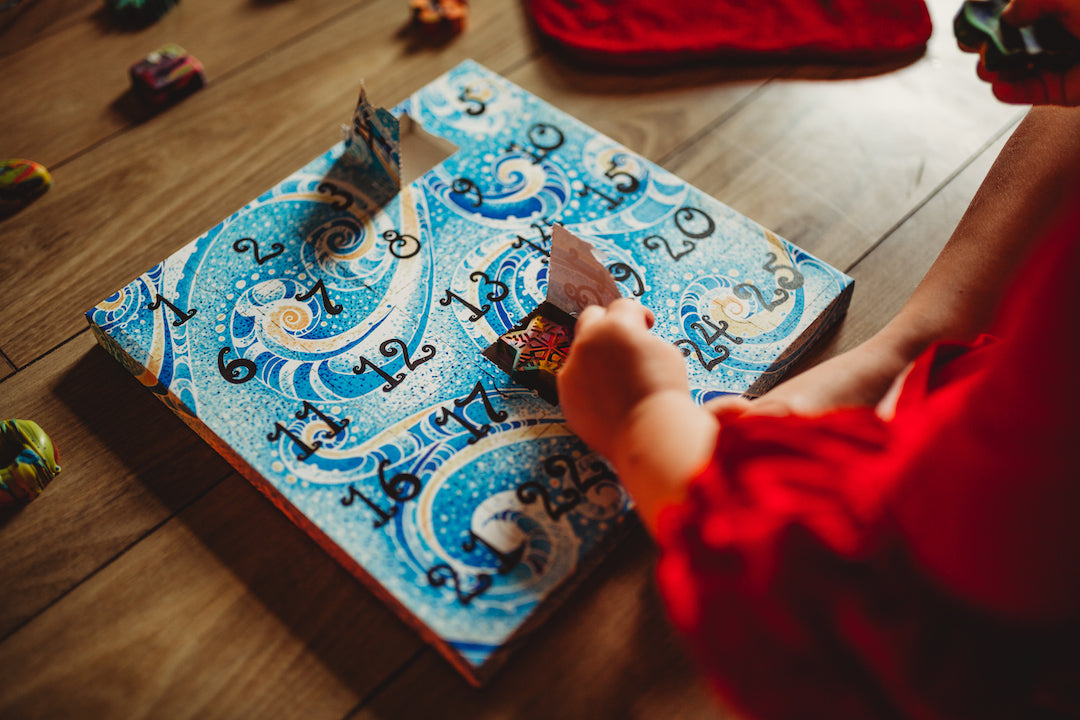Child in a red Christmas dress sits on a wooden floor with a blue crayon advent calendar. The child is opening paper door number 18 to reveal a snowflake rainbow crayon from Art 2 the Extreme. This Advent calendar for kids has a blue, teal, and light yellow swirl design and black numbering. 