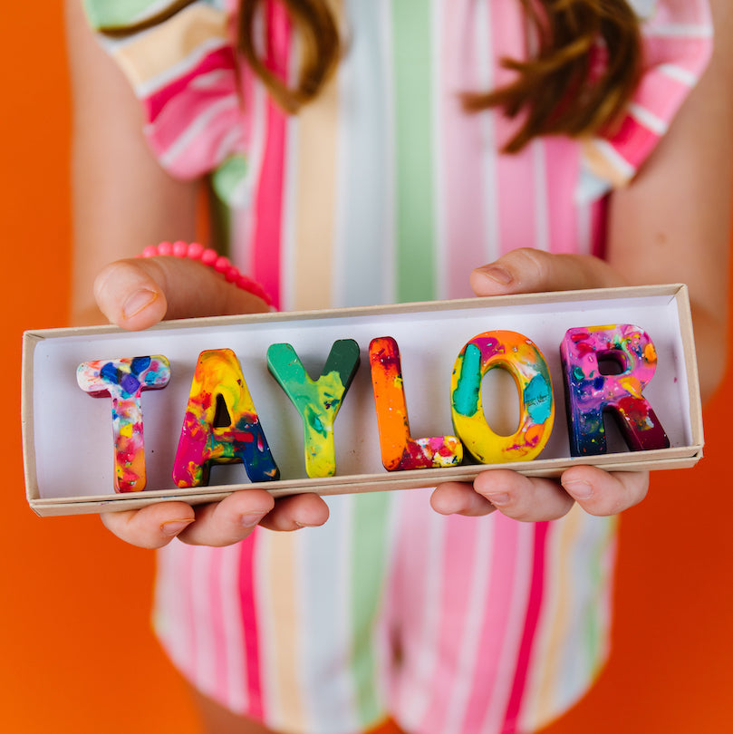A person in a pastel striped top holds a small box containing colorful crayon letters spelling "TAYLOR" against a bright orange background.