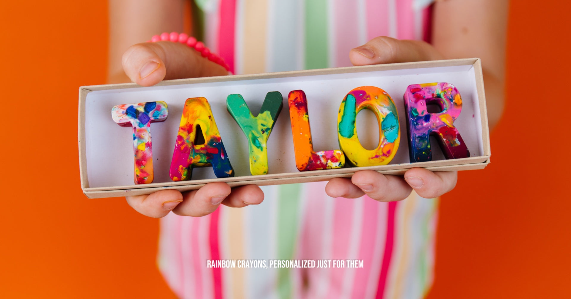 Child holding a boxed set of colorful personalized rainbow letter crayons spelling “TAYLOR,” with text reading “Rainbow Crayons, Personalized Just for Them.”