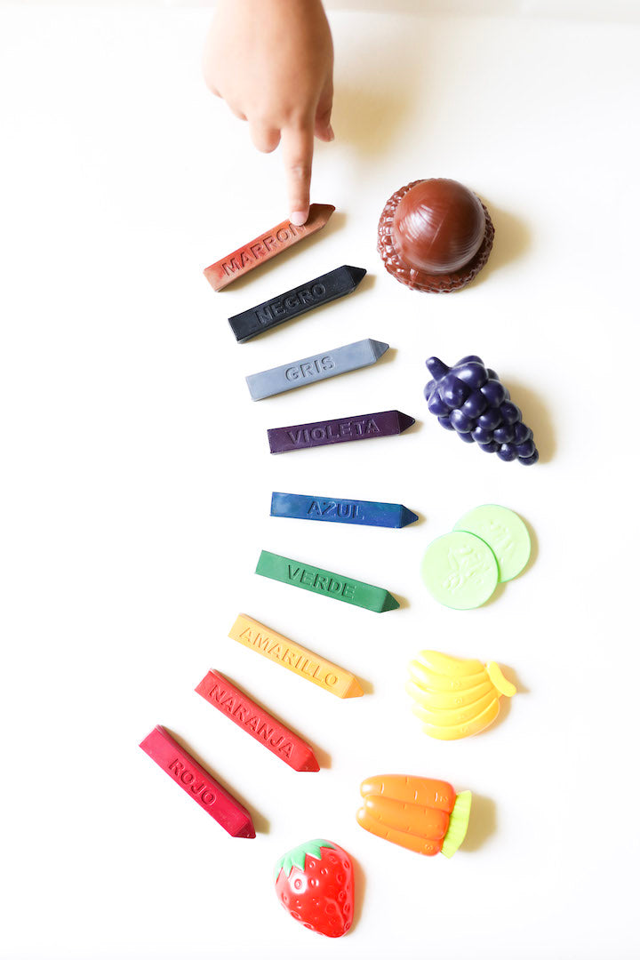 9 Solid colored crayons with Spanish names engraved on them sit on a white background with a small hand pointing at the Maroon (brown) crayon. There is a piece of plastic fruit of the same color next to each overused Crayon Stix®.